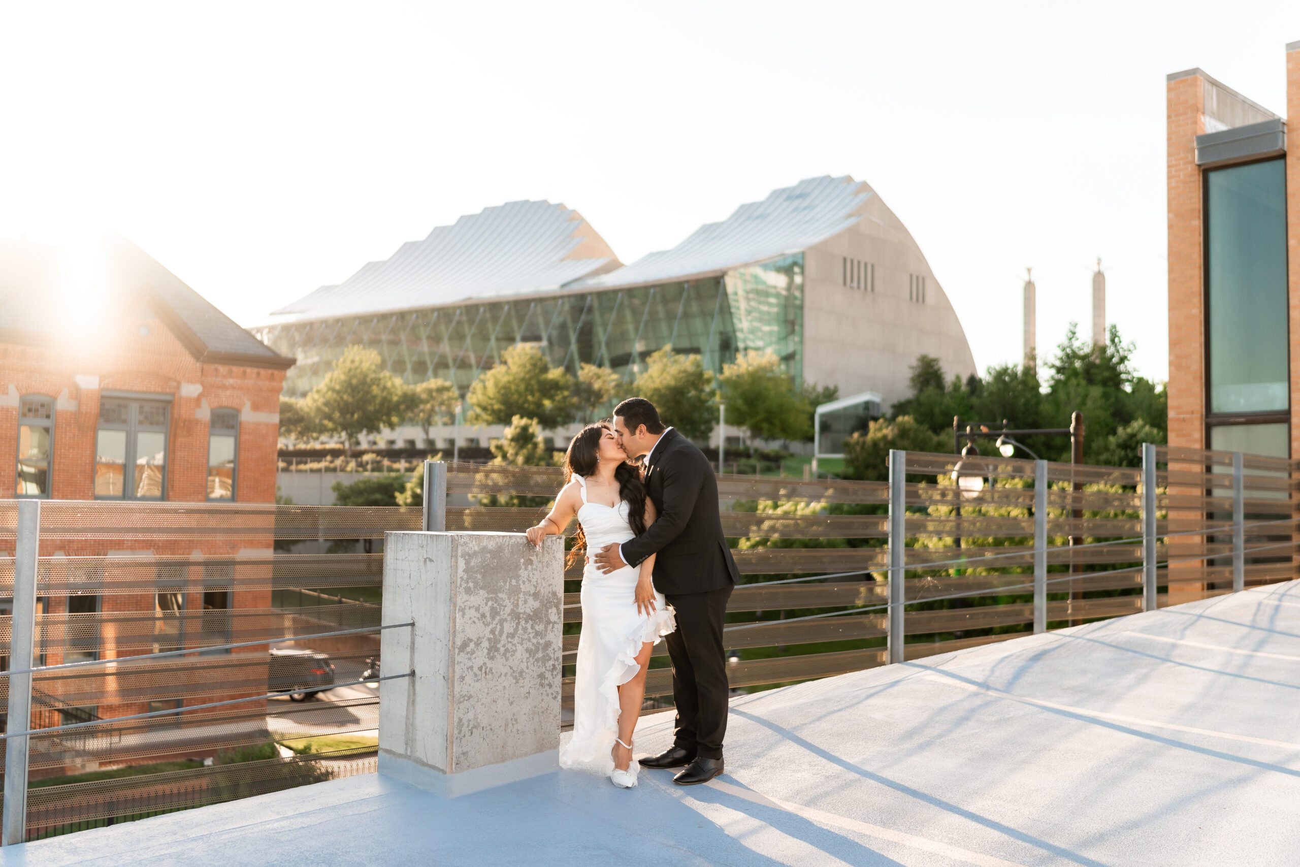 An engaged couple stands on a rooftop embracing each other and kissing. The woman is wearing a white dress and the man is wearing a black suit. The Kauffman Center can be seen in the background.