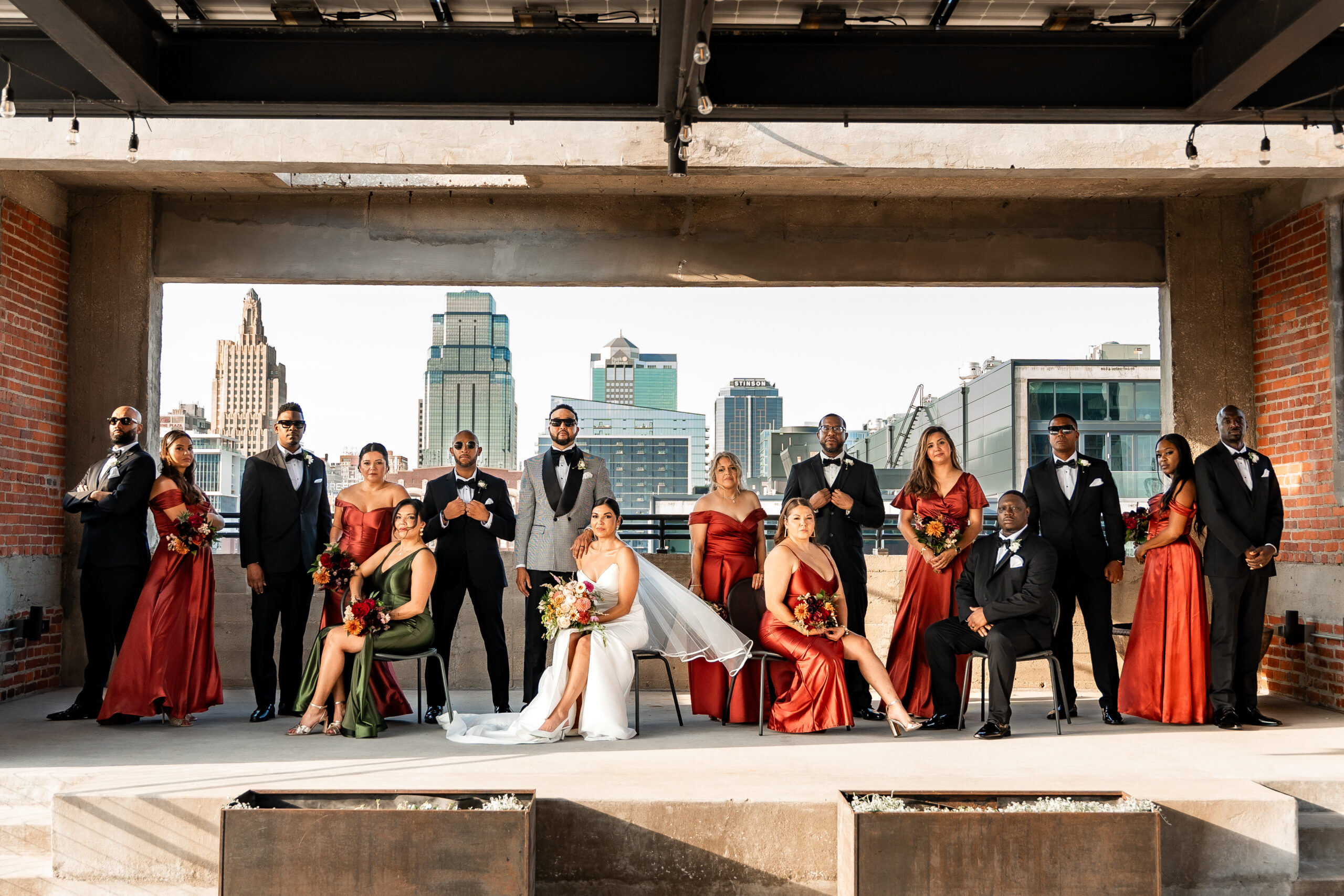 A bride and groom sit with their wedding party all around them, posed formally with the Kansas City skyline behind them