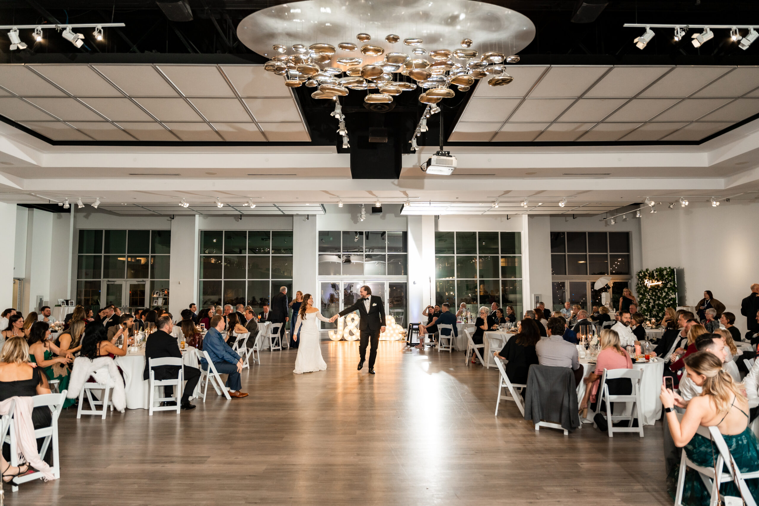 A bride and groom walk to the center of the Gallery Event Space dance floor surrounded by their seated friends and family